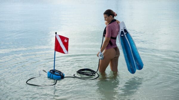 A woman standing in shallow water holding blue fins and a dive mask, with the BLU3 Nomad system floating next to her.