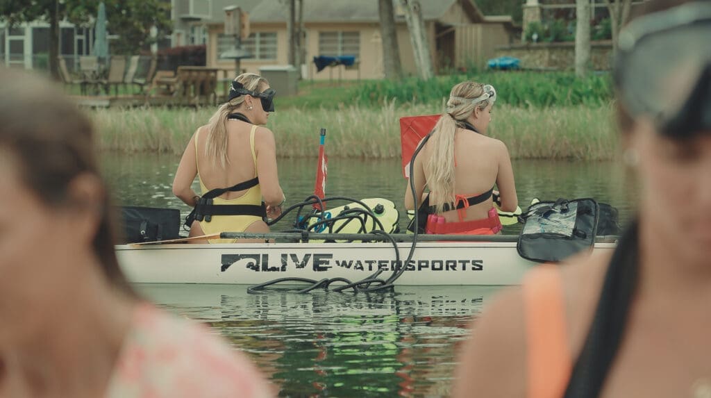 Two enthusiastic female divers are pictured on a Live Watersports paddle board, preparing for an underwater adventure. Clad in swimwear and equipped with BLU3 Nomad Dive Systems, they are meticulously checking their gear while sitting on the large, stable board, surrounded by clear turquoise waters. The joy of anticipation for their dive is palpable in the air, perfectly encapsulating the spirit of water sports and exploration