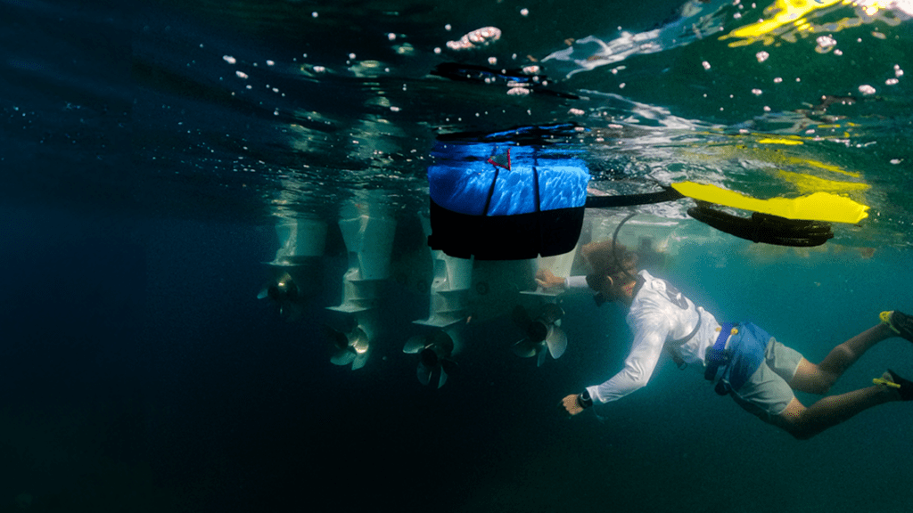 A diver using the BLU3 Nomad Mini dive system for a boat hull inspection, connected to a floating blue dive unit. The diver is seen swimming under a boat with visible propellers.