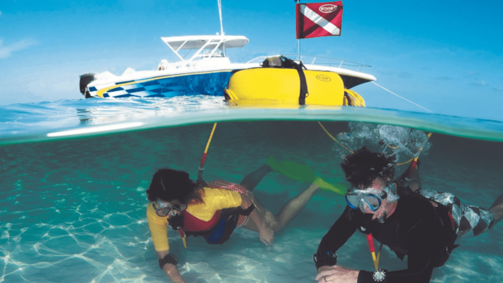 Two divers using Brownie's Third Lung surface-supplied air system in shallow water, connected to a yellow floating unit with a boat and dive flag in the background.