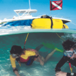Two divers using Brownie's Third Lung surface-supplied air system in shallow water, connected to a yellow floating unit with a boat and dive flag in the background.
