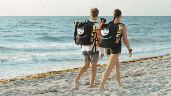 Two people walk down the beach with BLU3 branded backpacks on with dive systems packed inside the backpacks showing the portability of BLU3 dive systems.