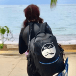 Underwater photographer Kate UA walking towards the beach carrying her BLU3 Nomad dive system backpack, dressed in a wetsuit, ready for a dive.