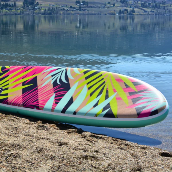 10'6 Royal Hawaiian Palm inflatable paddle board resting on a lakeside shore, showcasing its vibrant tropical palm design