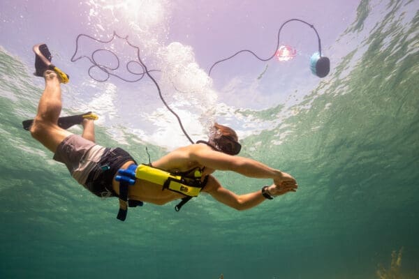 Diver in the ocean swimming underwater showing the Nomad floating above him with the hose clearly connected from the BLU3 Nomad battery powered dive system down to the harness of the diver for breathing without scuba tanks.