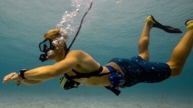 Diver using a BLU3 tankless dive system with surface-supplied air hose and a Spare Air emergency backup tank mounted on harness in shallow water.