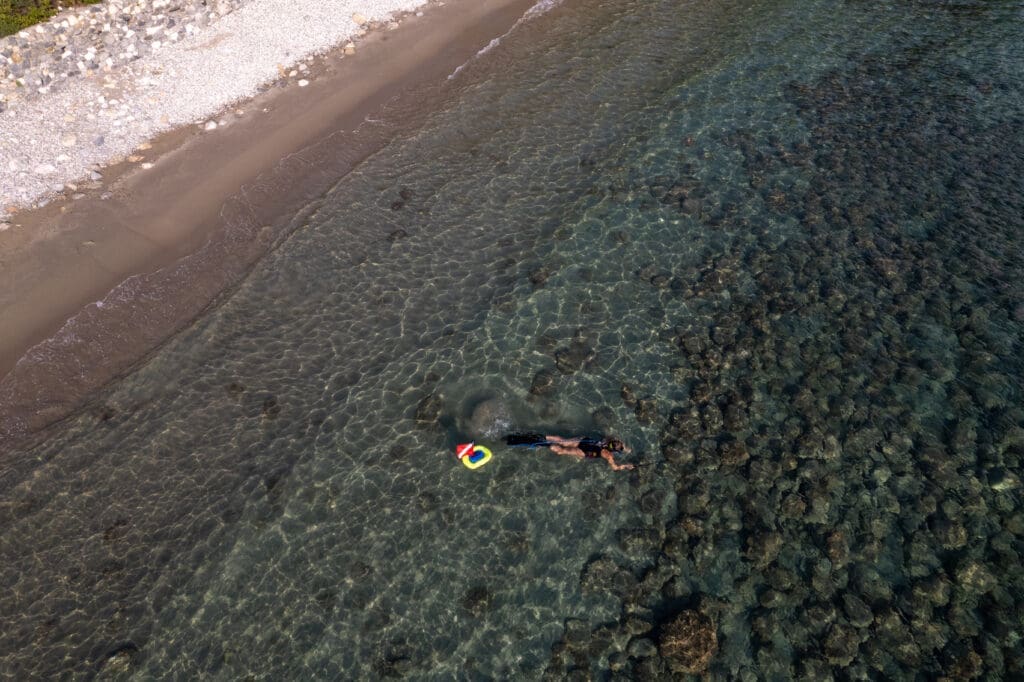 Aerial view of a diver using a BLU3 tankless dive system with a floating dive flag near a rocky shoreline in shallow water.