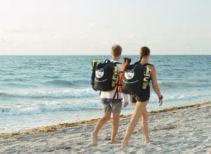 Two people walking barefoot along the beach carrying BLU3 Nomad Mini tankless dive systems in backpacks, heading toward the ocean for a shallow diving session.